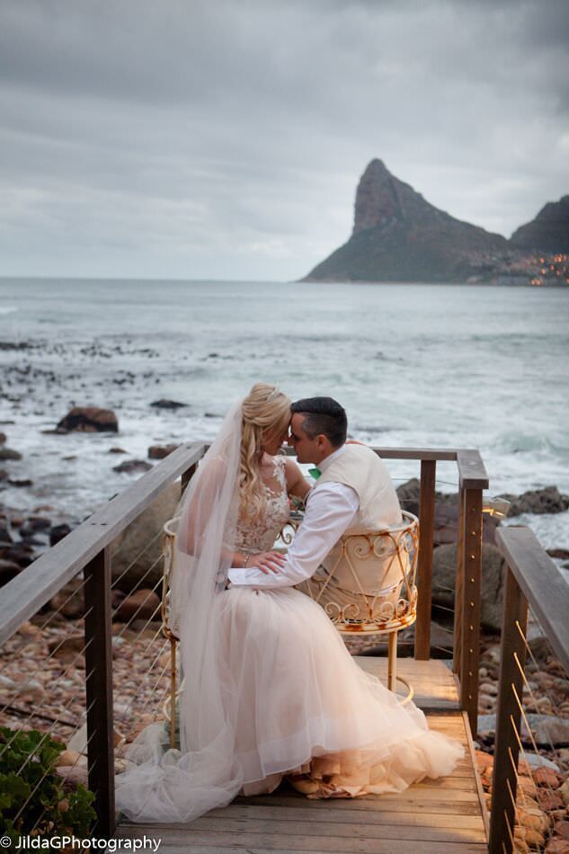 A bride and groom are kissing on a wooden bridge overlooking the ocean.