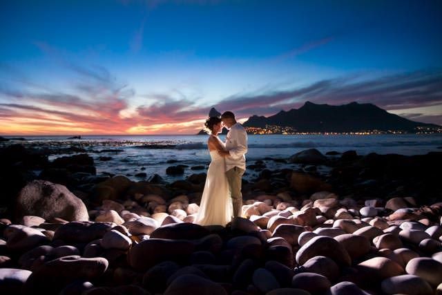 A bride and groom are kissing on a rocky beach at sunset.