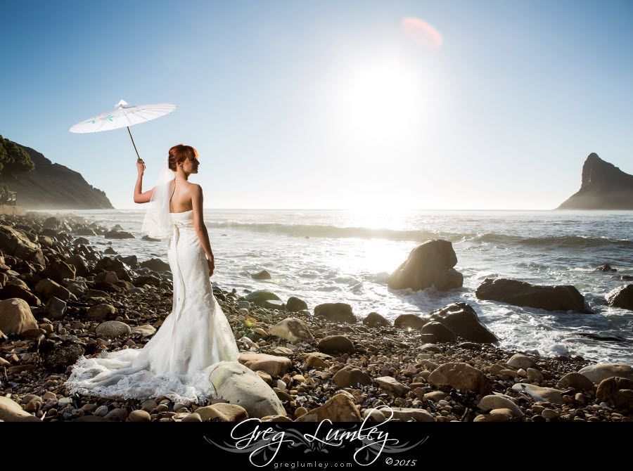 A bride in a wedding dress is holding an umbrella on the beach.