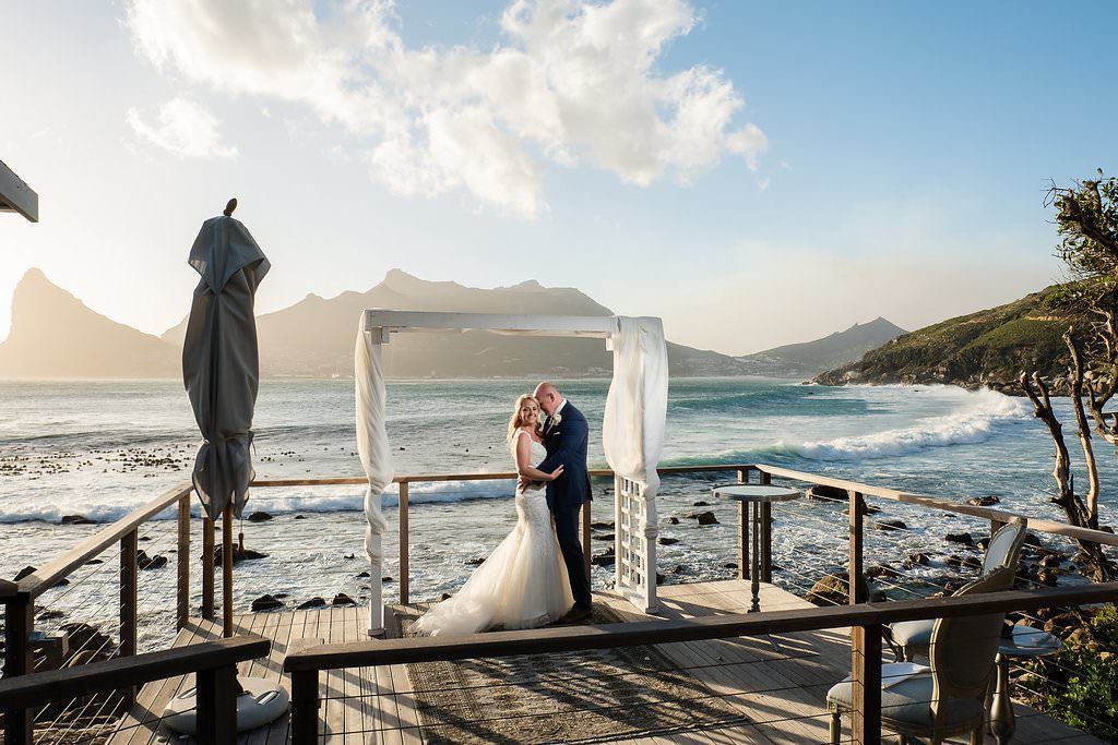 A bride and groom are kissing on a pier overlooking the ocean.