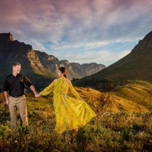 A man and a woman are holding hands in a field with mountains in the background.
