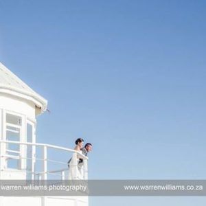 A bride and groom are standing on a balcony overlooking the ocean.