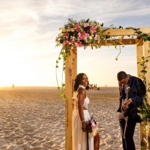 A bride and groom are standing under a wooden arch on the beach.