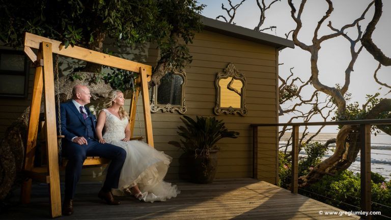 A bride and groom are sitting on a wooden swing in front of a house.
