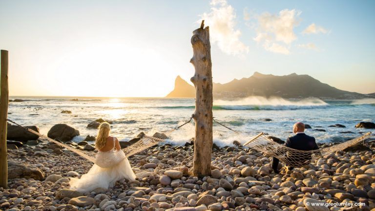 A bride and groom are sitting in hammocks on the beach at sunset.