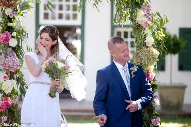 A bride and groom are standing under a floral arch at their wedding.
