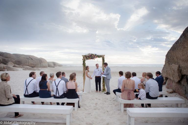 A group of people are sitting on the beach watching a wedding ceremony.