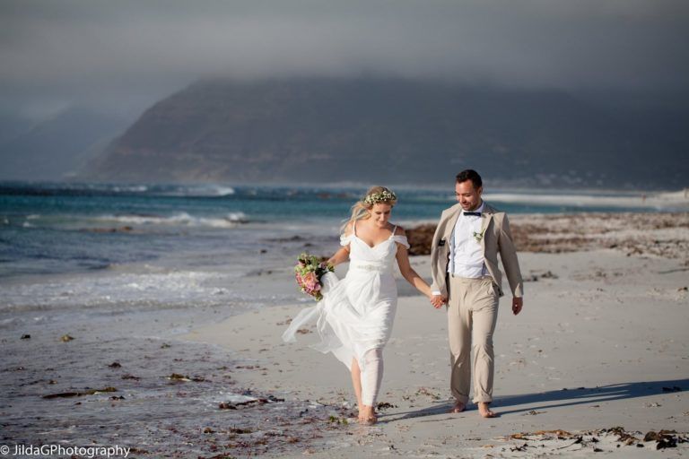A bride and groom are walking on the beach holding hands.