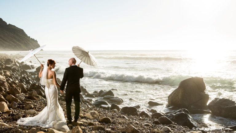 A bride and groom are holding umbrellas on the beach.