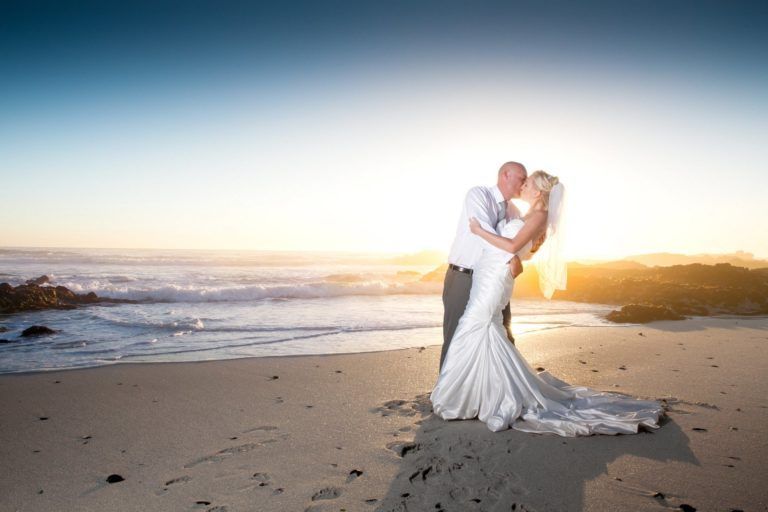 A bride and groom are kissing on the beach at sunset.