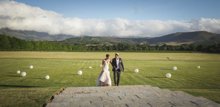 A bride and groom are standing in a field with mountains in the background.