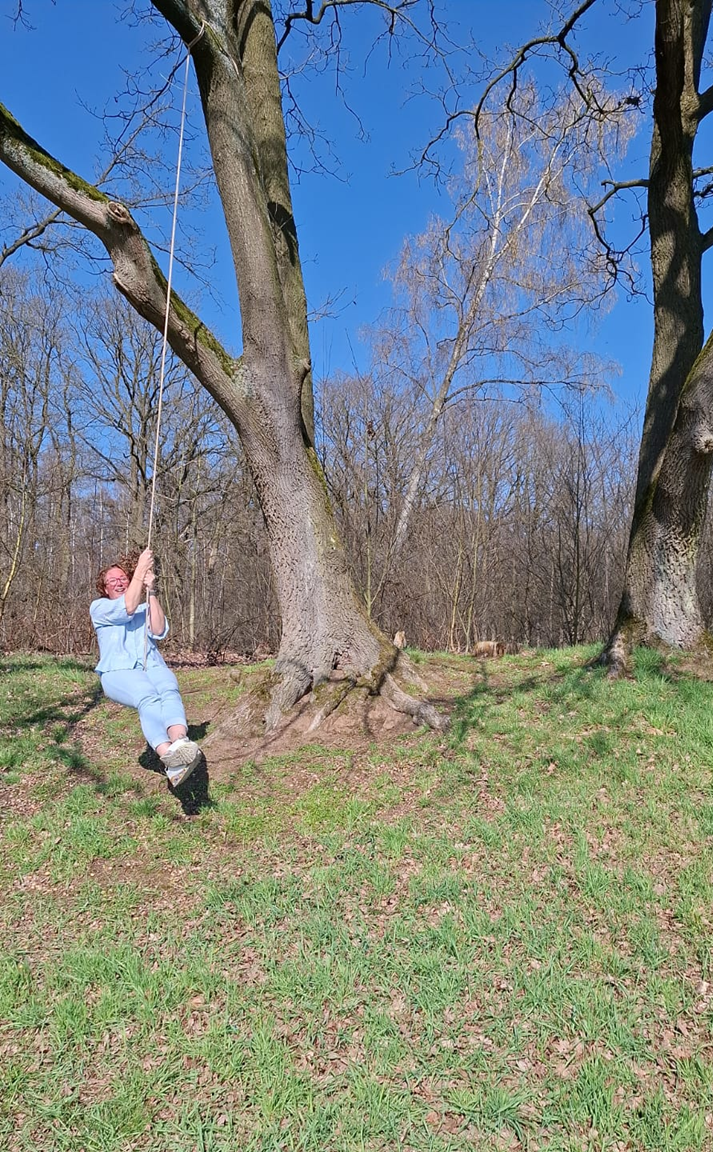 Een persoon in lichtblauwe kleding slingert aan een touw dat aan een grote boom in een grasveld is bevestigd.