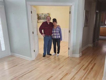 Couple standing in doorway, facing camera. Room with light-colored hardwood floors, framed doorway, and light blue walls.