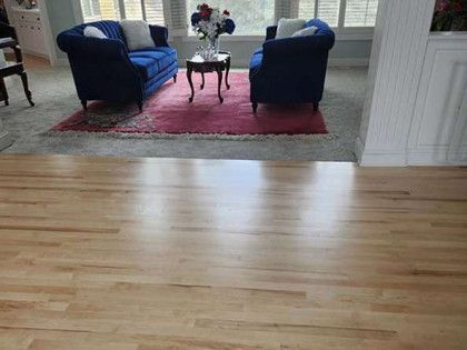 Hardwood floor in foreground, leads to a living room with blue sofas, a rug, and windows.