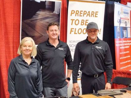 Three people stand at a trade show booth with flooring samples.