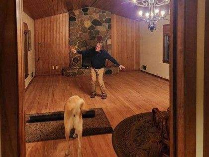 Man dancing in empty room with dog, wood paneling, fireplace, chandelier, and area rugs.
