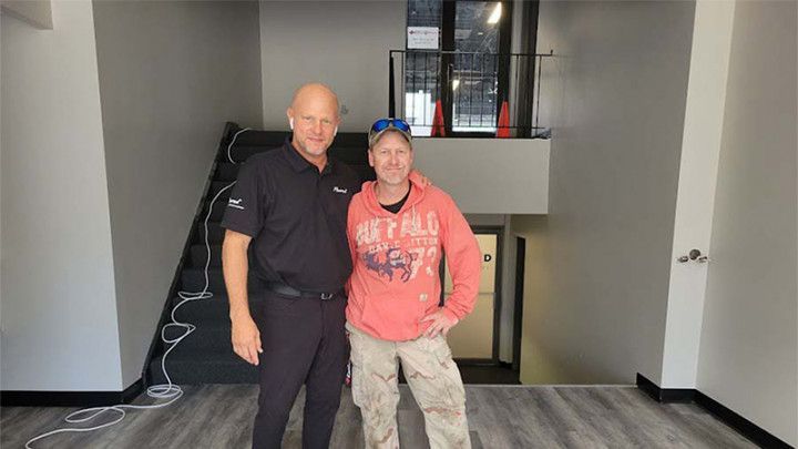 Two men standing in a renovated interior space, a staircase in the background, one in black shirt, the other in red sweatshirt.