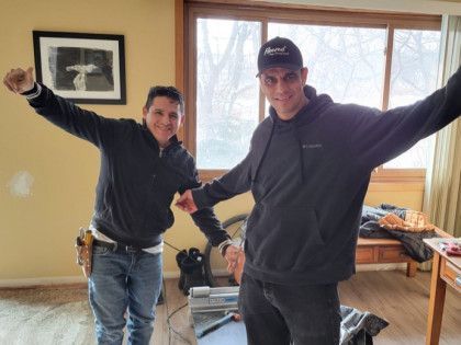 Two men with arms outstretched in a room, one holding a tool belt. Beige walls, window in background.