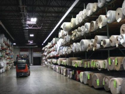 Warehouse interior with rolls of carpet on shelves, a forklift, and loading dock.