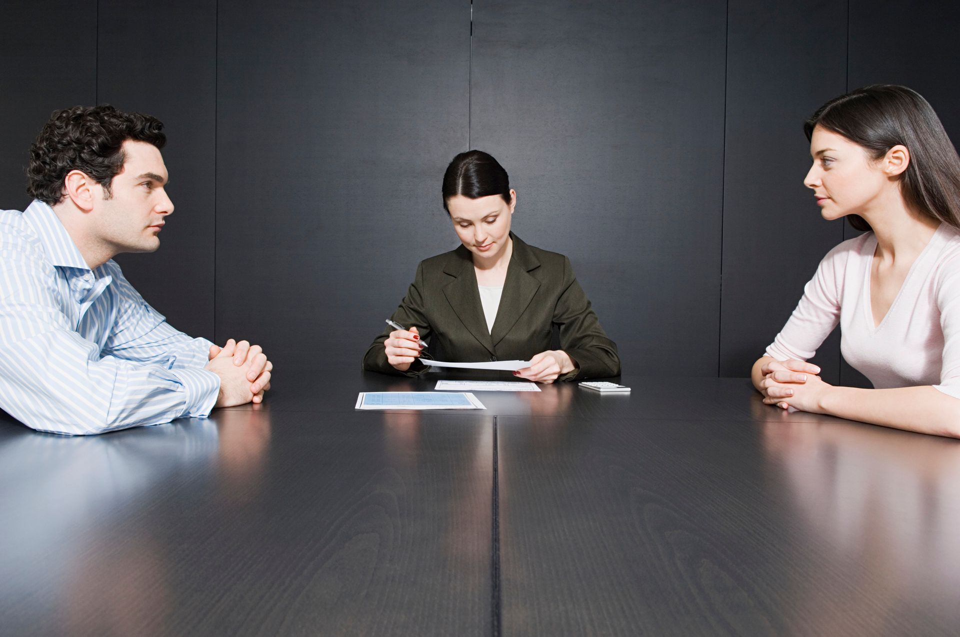 Couple at a table, facing a person in a suit, possibly a mediator or lawyer; serious expressions.