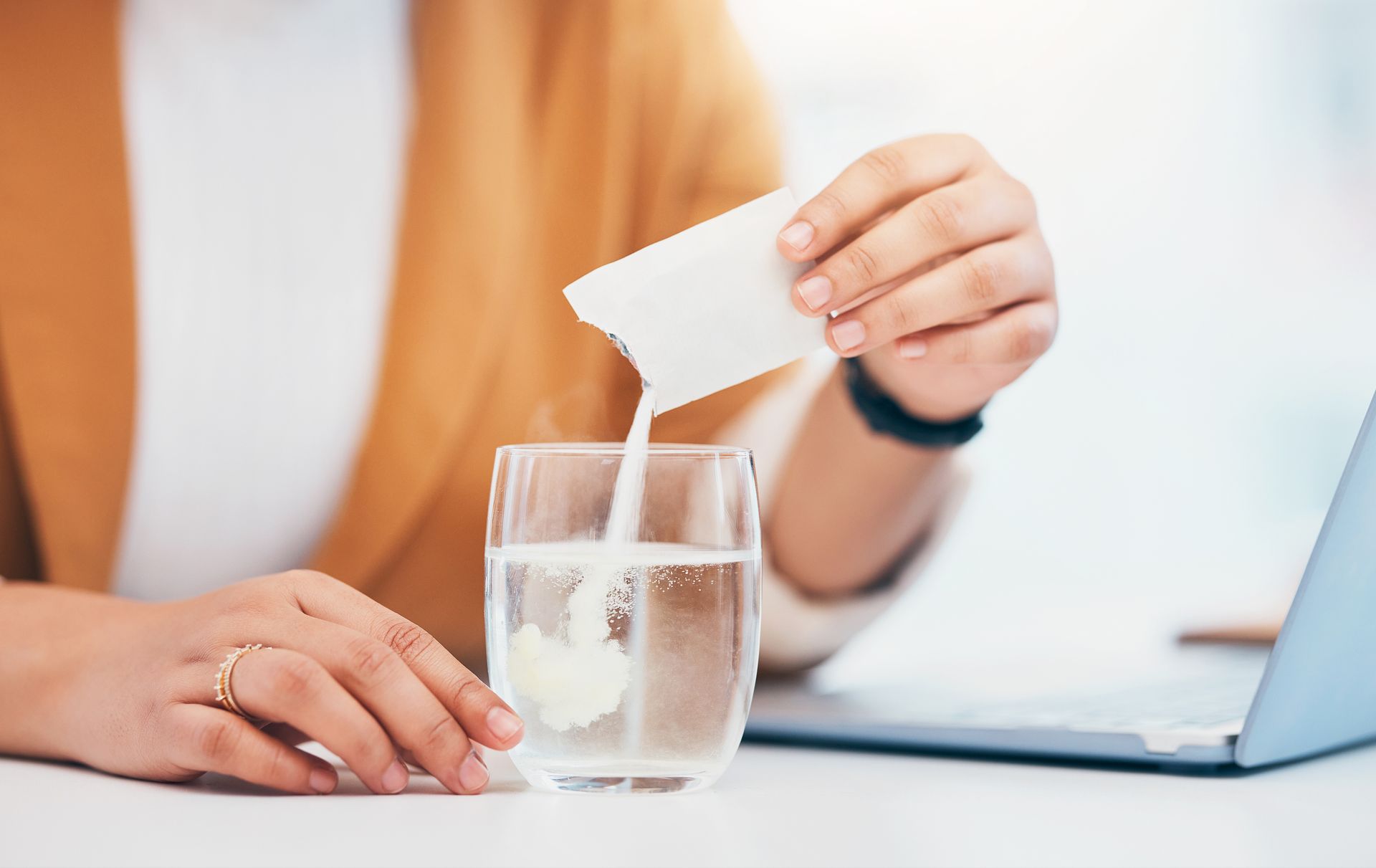 Person pouring powder from a packet into a glass of water, near a laptop Person pouring powder from a packet into a glass of water, near a laptop