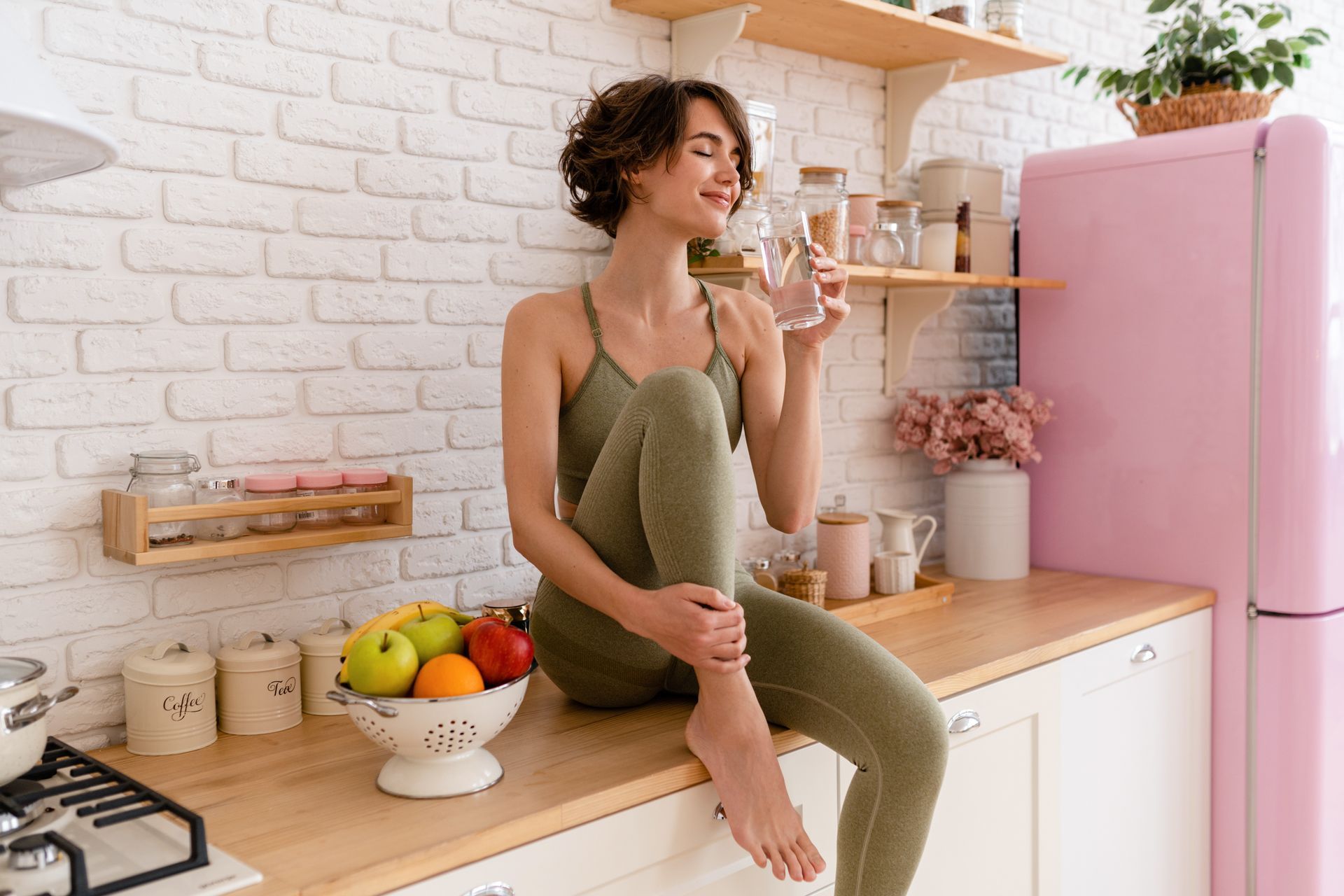 Woman in green activewear drinks water in a kitchen with fruit
