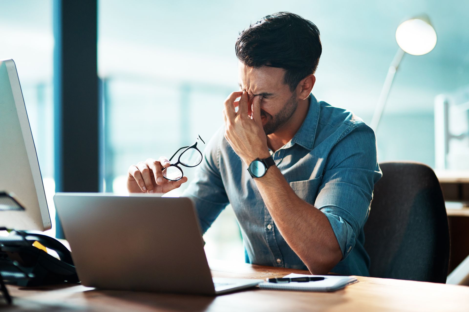 Man in blue shirt rubbing eyes, holding glasses, at a laptop in office