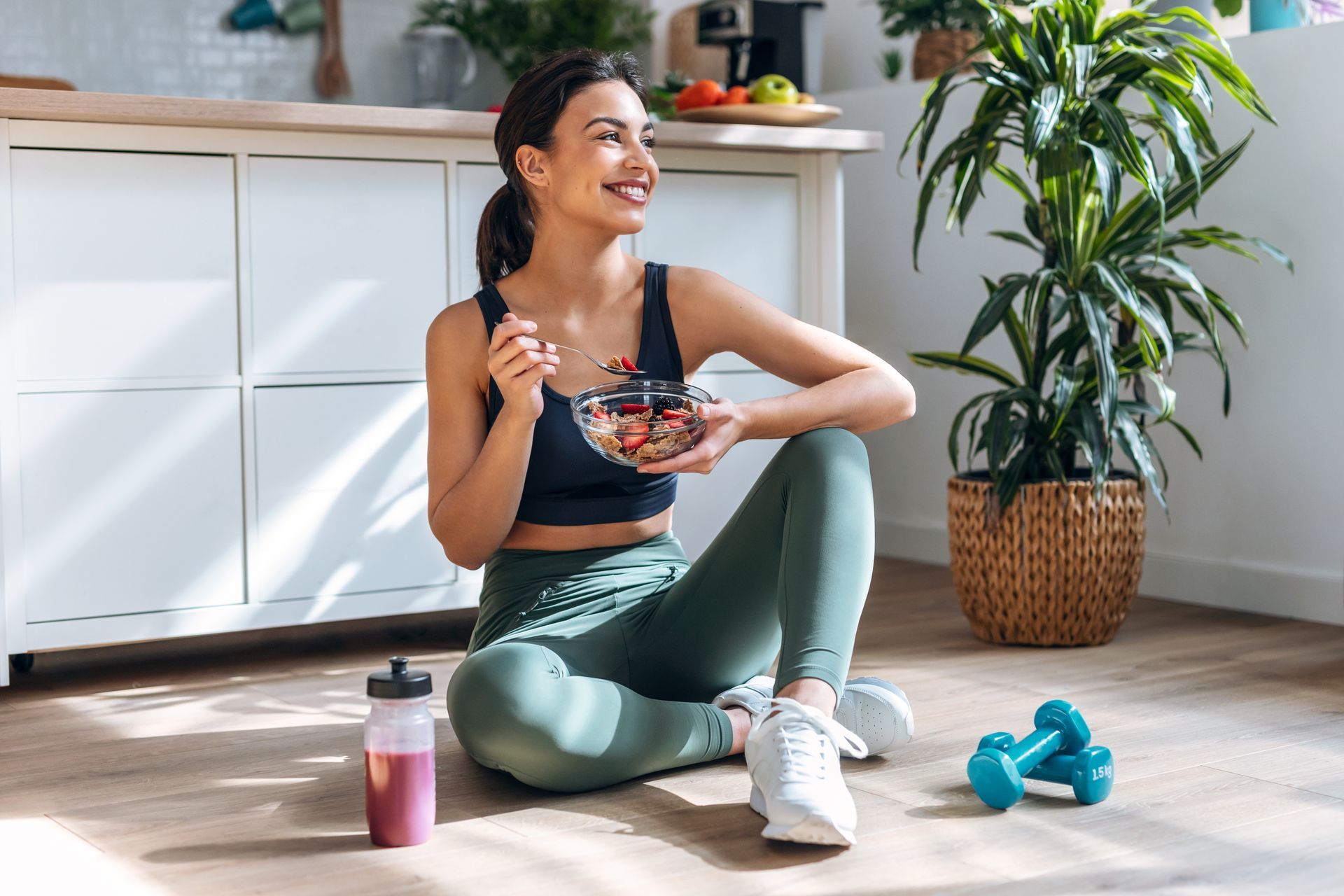 Woman in athletic wear sitting, eating fruit, with dumbbells and water bottle nearby