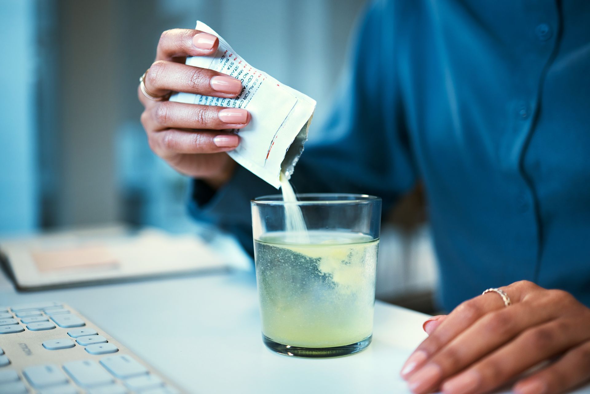 Person pouring powder from a packet into a glass of water, next to a computer keyboard Person pouring powder from a packet into a glass of water, next to a computer keyboard