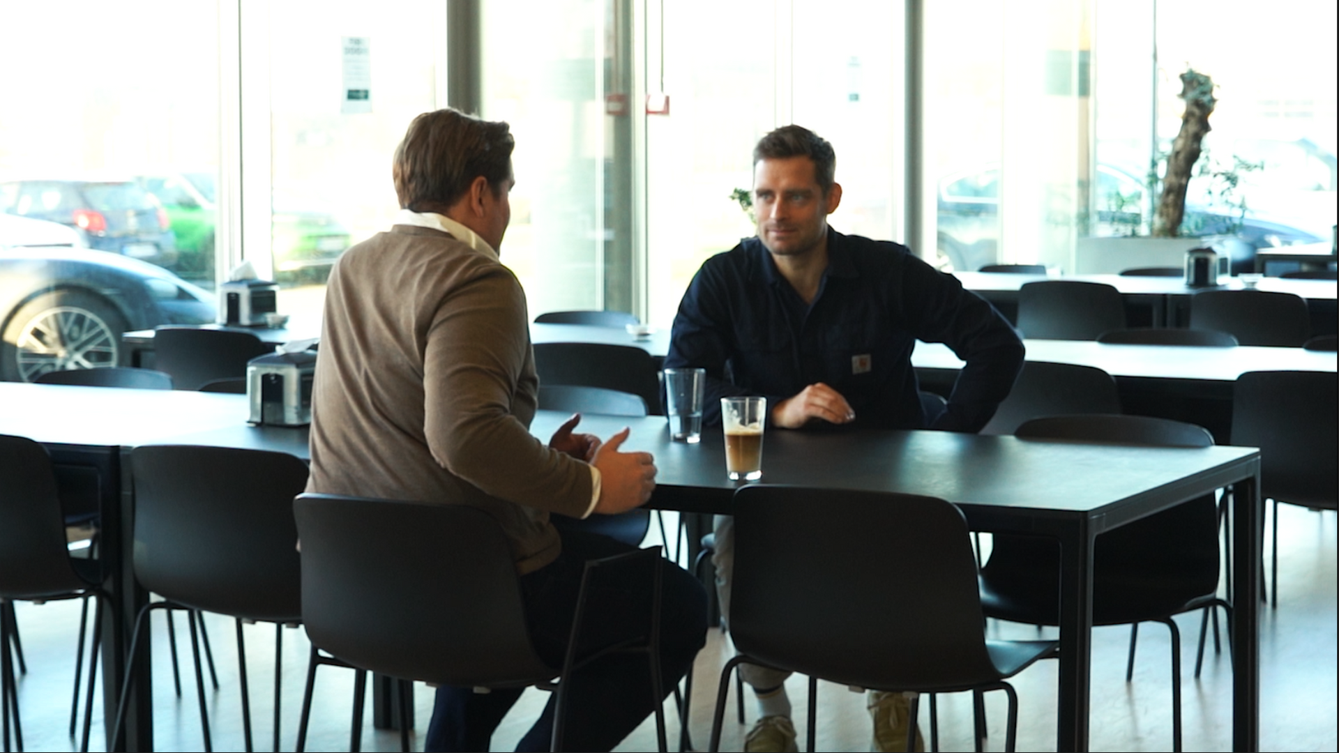 Two men seated at a table in a modern, brightly lit cafe, engaged in a conversation.