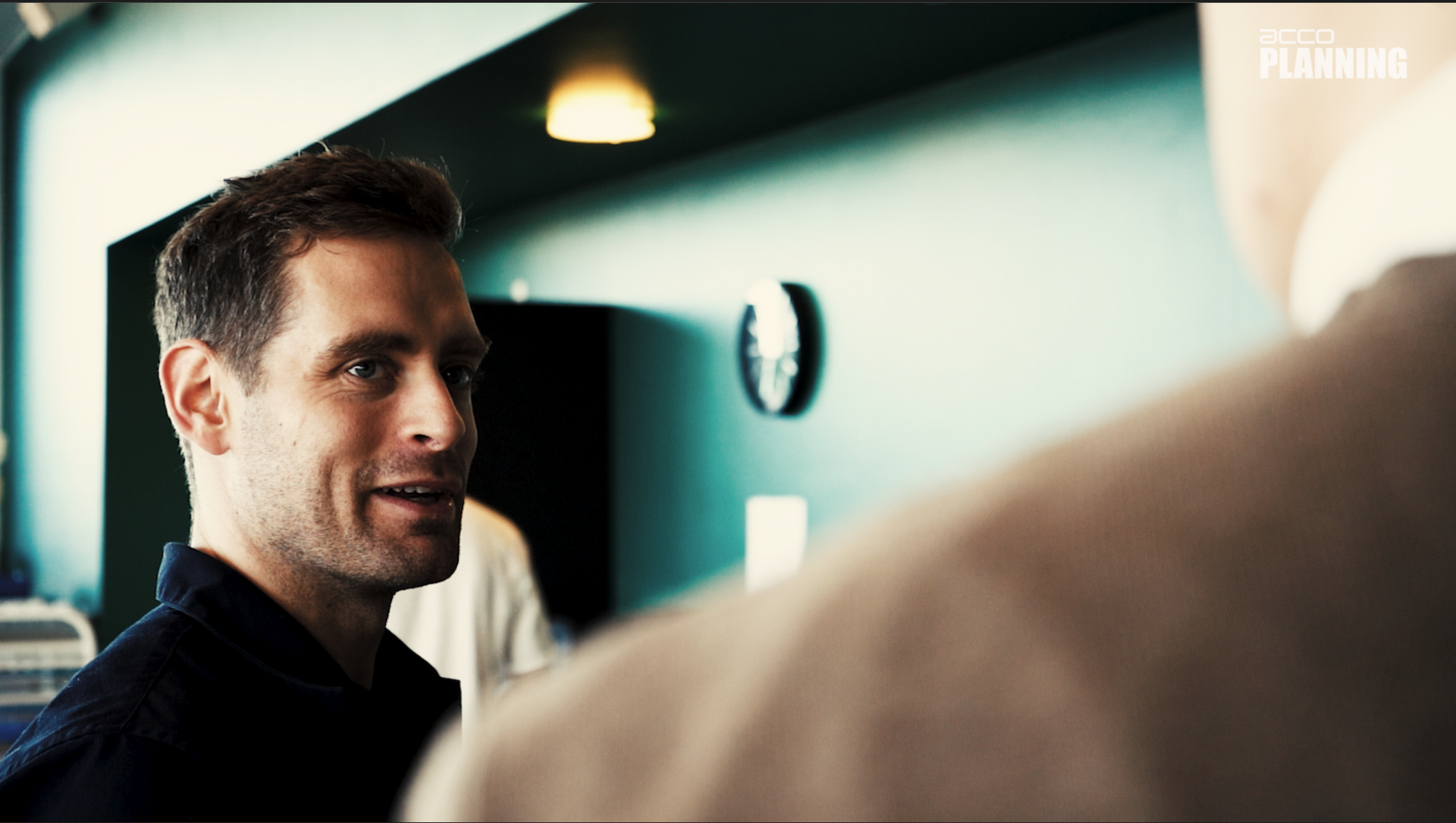 Man smiles, facing right, in a teal-walled room. Another man is out of focus in the foreground, wearing a tan coat.