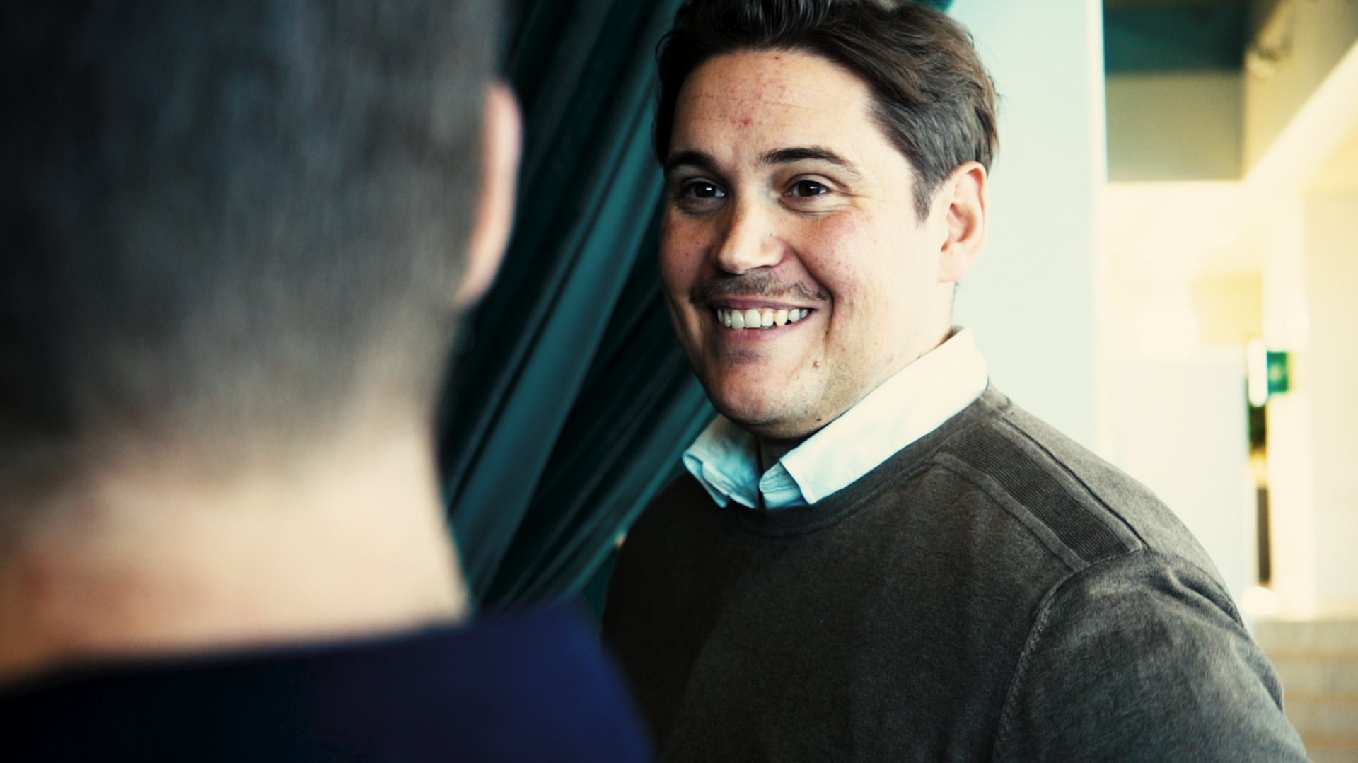 Man smiling, facing another person. He's wearing a sweater and button-down shirt in a brightly lit room.