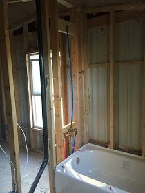 Bathroom under construction with tub installed, plumbing visible in wood frame next to a window.