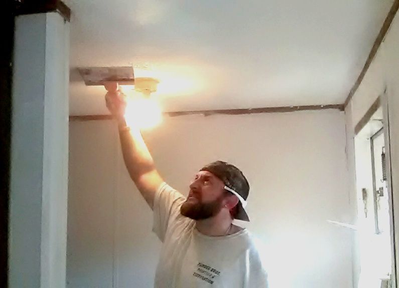 Man using a drywall knife on a ceiling. He is wearing a cap and face mask, indoors, lit by a bright light.