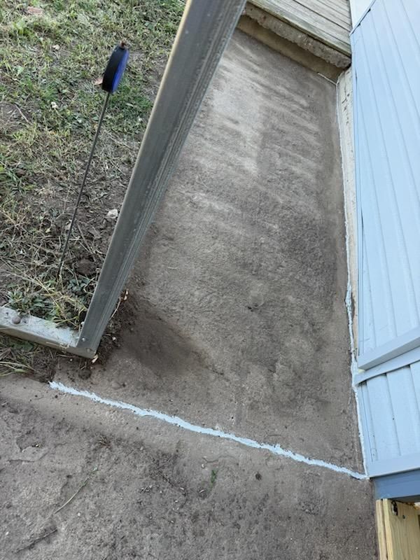 Gray concrete patio next to a house and under a wooden structure; white caulk is visible.