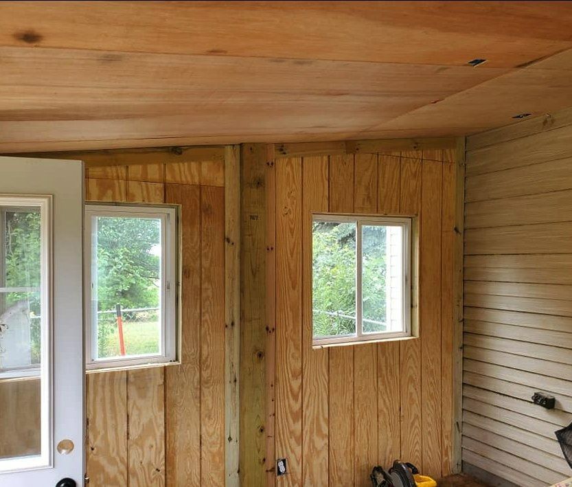 Interior of a room under construction with plywood walls and ceiling, two windows, and a door frame.