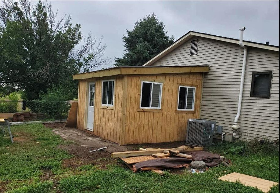 A small, light-brown building attached to a beige house, with windows, a door, and construction materials scattered nearby.