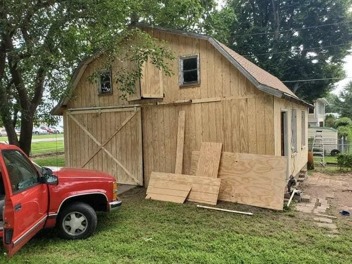Red truck parked near a wooden barn under construction, surrounded by grass and trees.