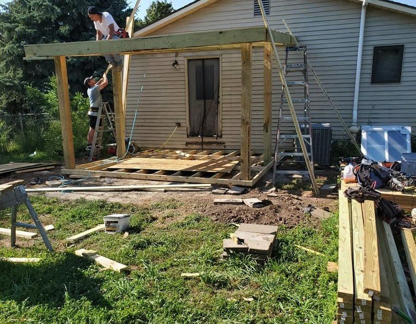 Two people building a wooden patio cover attached to a house with clapboard siding; tools and materials scattered.