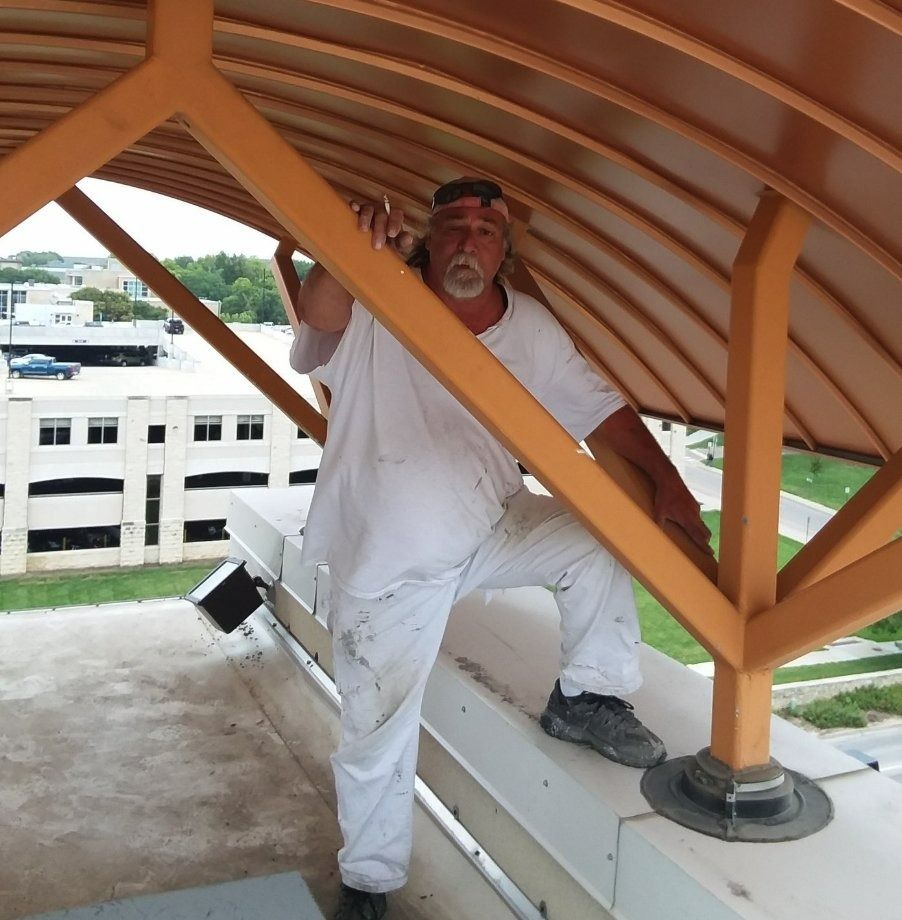 Man in white work clothes stands on a roof, holding onto a brown beam.