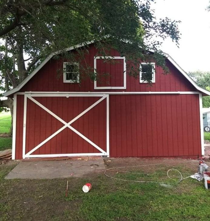 Red barn with white trim, door, and an X design. Concrete pad in front, grass and tree in background.