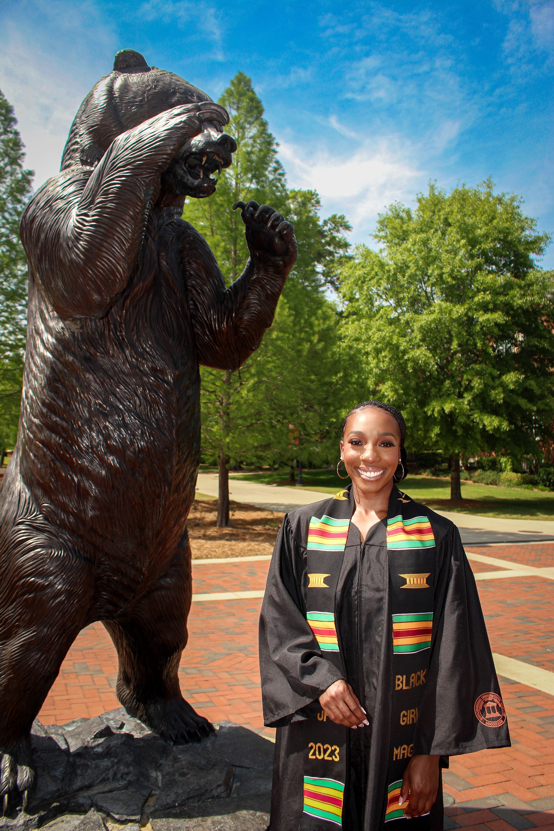 Person standing beside a large bear statue in a park, wearing a black coat with colorful trim.