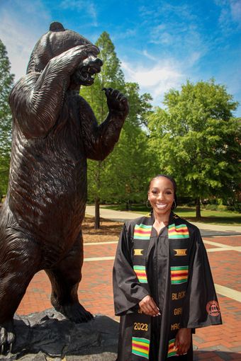 Person standing beside a large bear statue in a park, wearing a black coat with colorful trim.