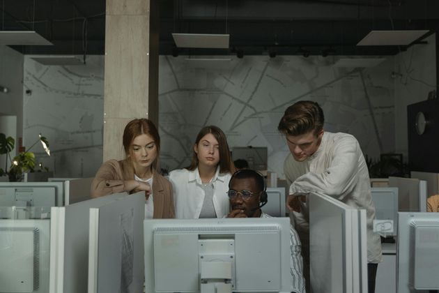 Four coworkers gathered around computer monitors in a dim office, reviewing something together