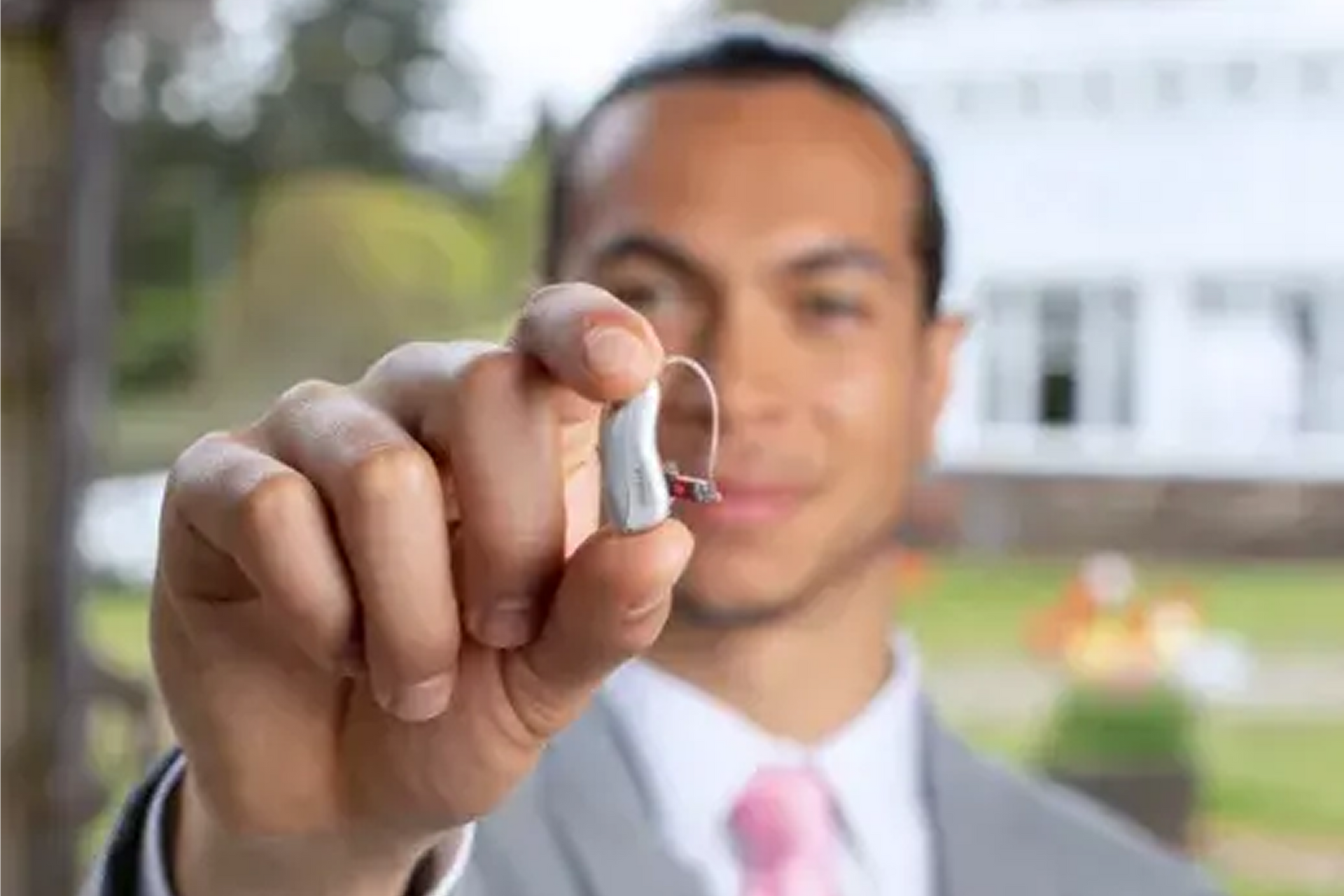 Man in suit holds up a hearing aid; soft focus background of a building and greenery.