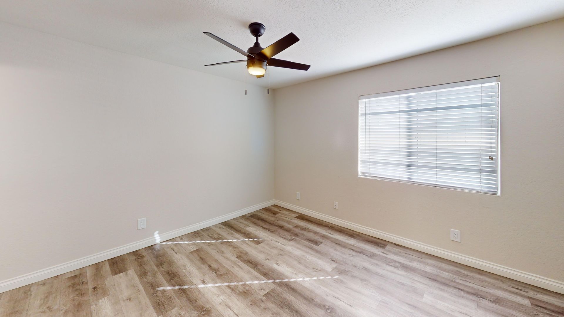 Empty bedroom with gray wood-look floor, window with blinds, and ceiling fan. Beige walls.