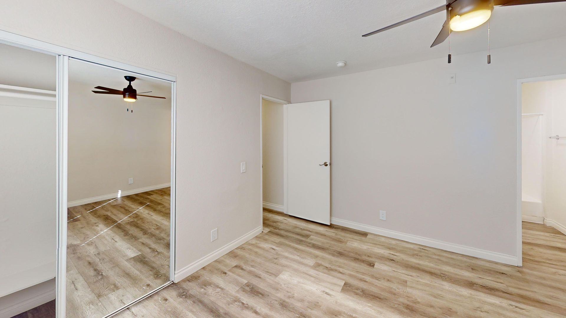 Bedroom interior with light wood flooring, white walls, mirrored closet doors, and ceiling fan.