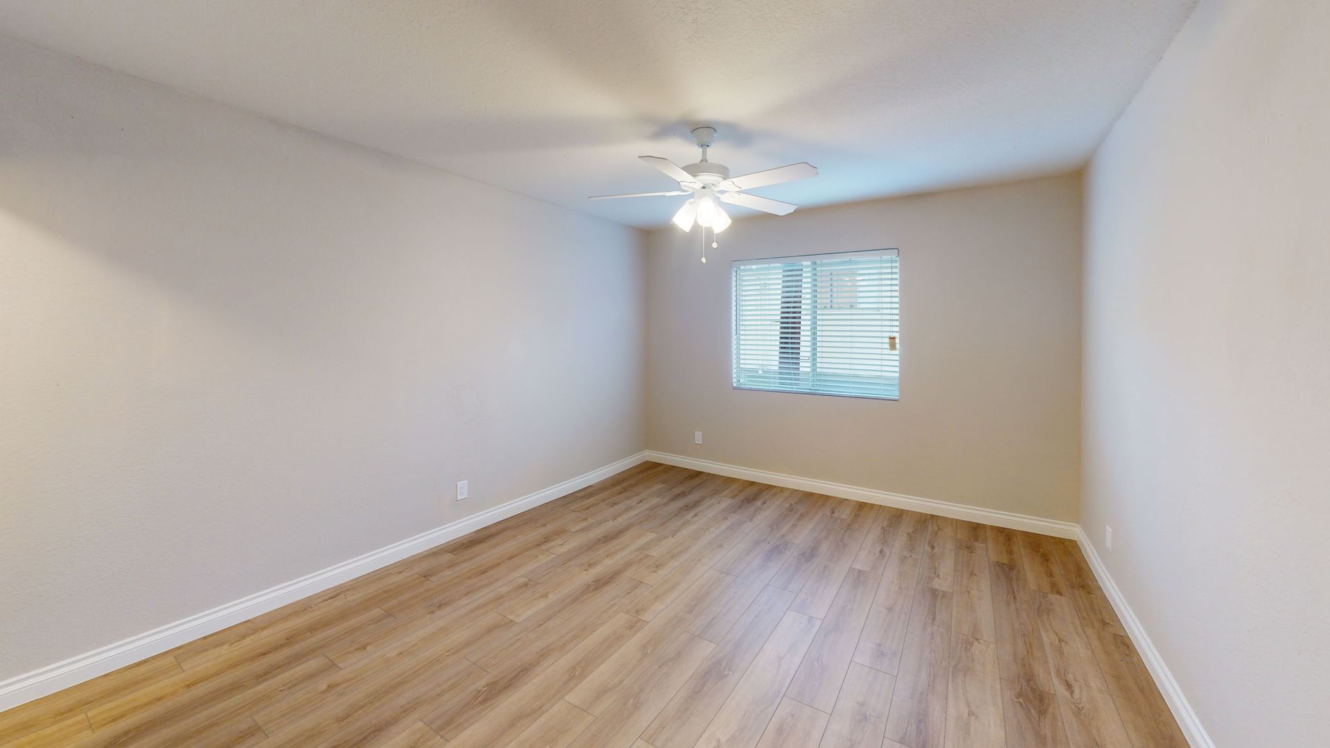 Empty bedroom with wood-look floor, window, and ceiling fan. Beige walls and white trim.