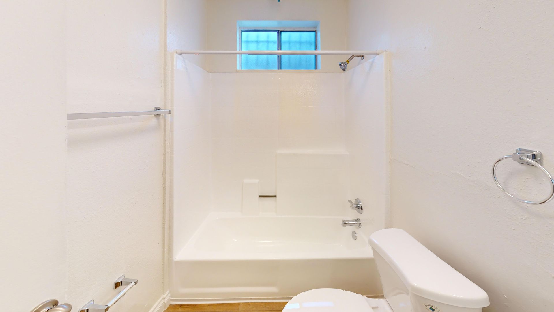 White bathroom with shower, toilet, and towel rack. Sunlight through a small window above the shower.