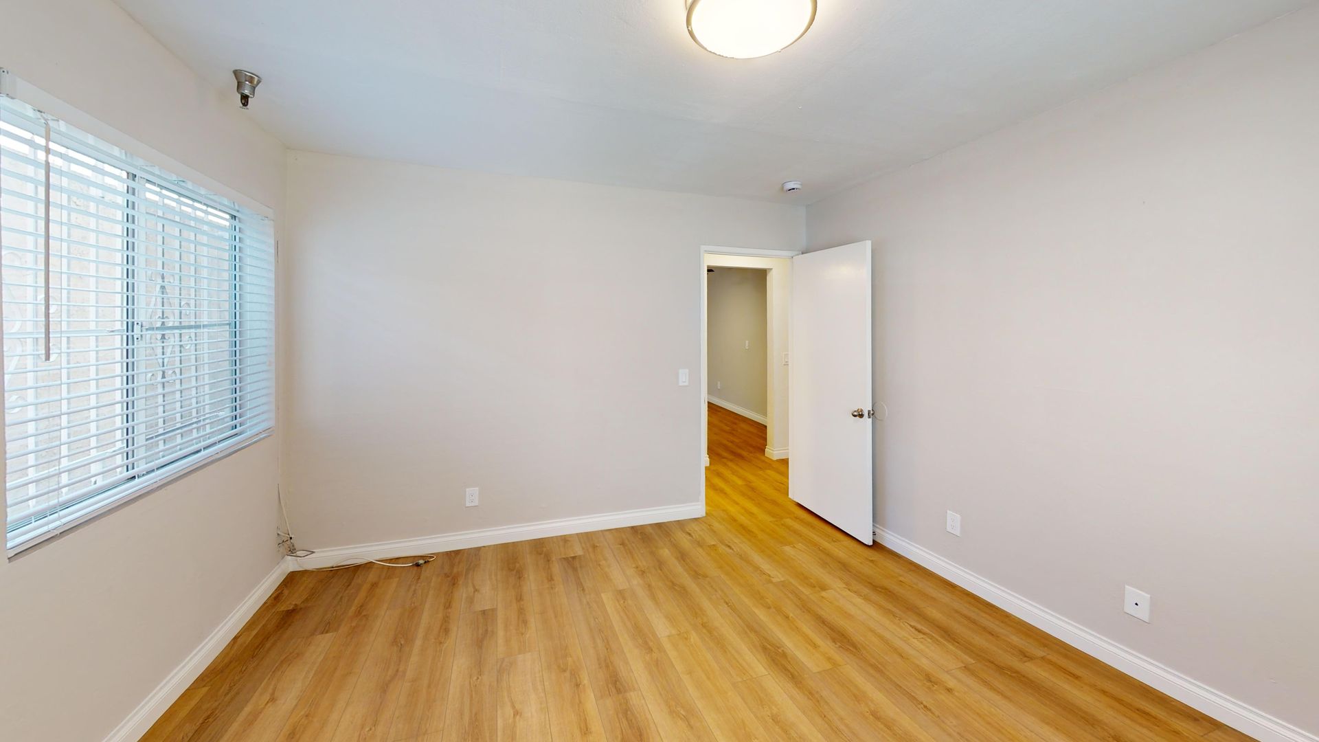 Empty room with wood floors, a window with blinds, and a door leading to a hallway.